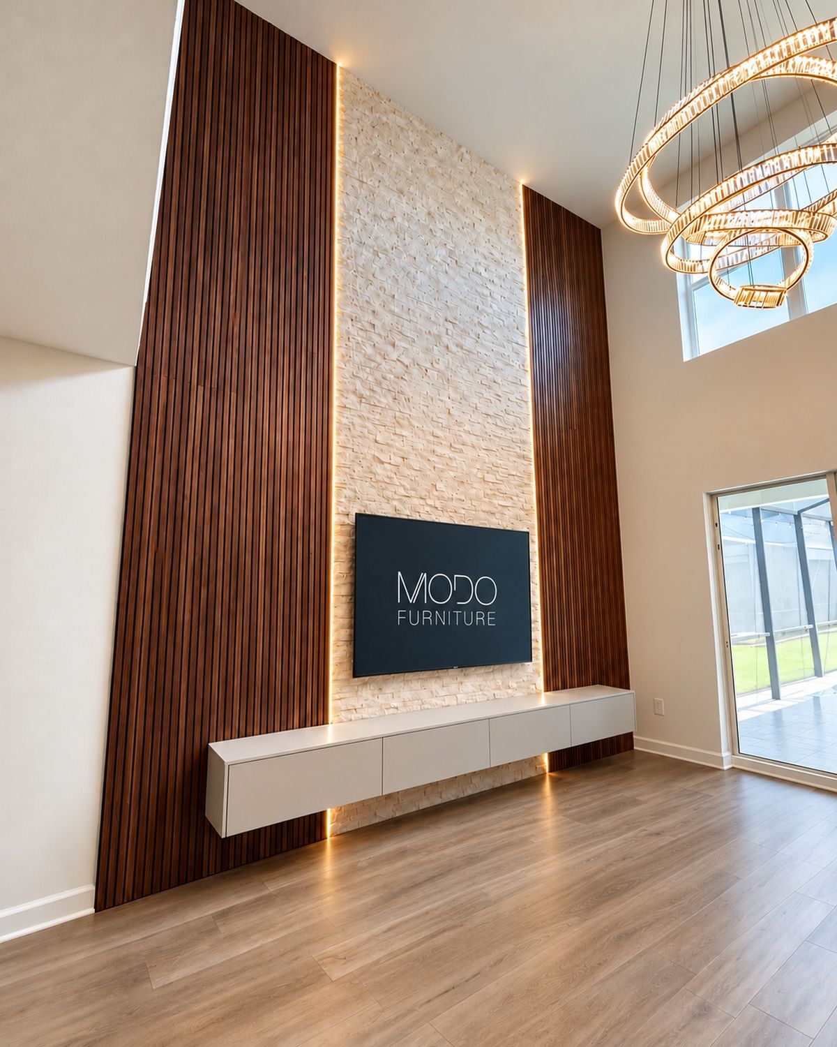 Floating white TV console below a TV wall with tall walnut slat panels and backlit stacked stone, with a chandelier above