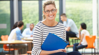 A female teacher smiling to the camera with a classroom in the background.