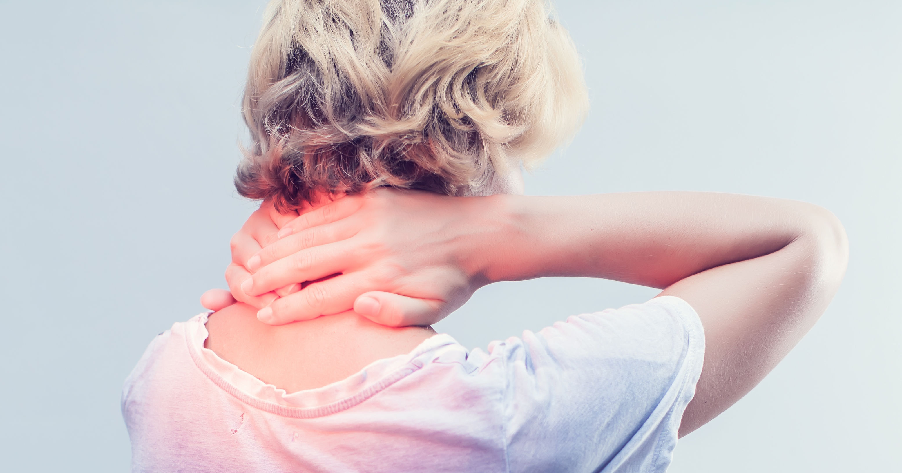 A person with short hair is seen from behind, holding their neck in discomfort while wearing a loose-fitting white shirt against a light background.