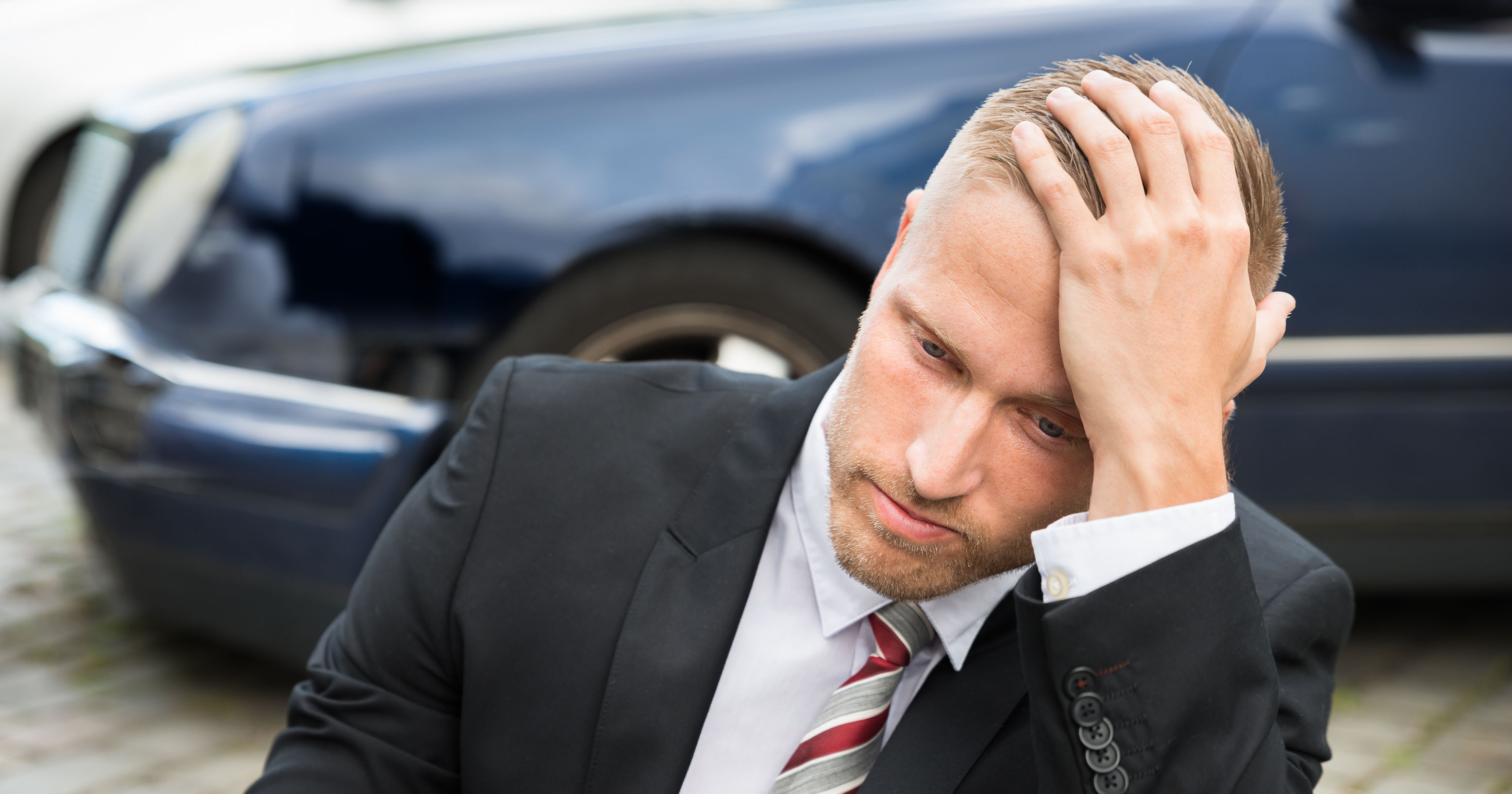 Young man in suit with hand on head sits on ground in front of wrecked car
