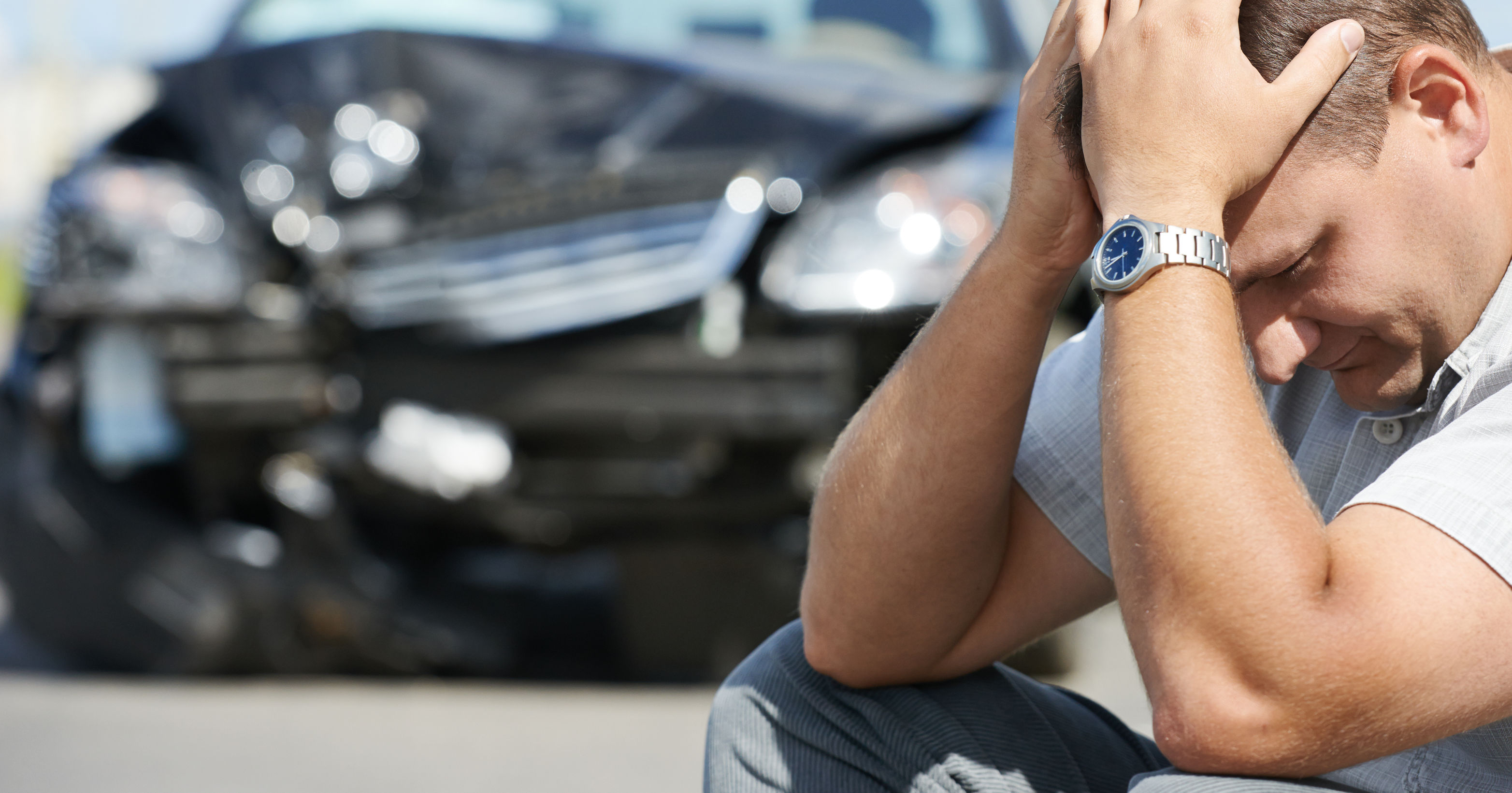 A man sits on the ground looking distressed, holding his head, with a damaged black car in the background, indicating a recent accident.