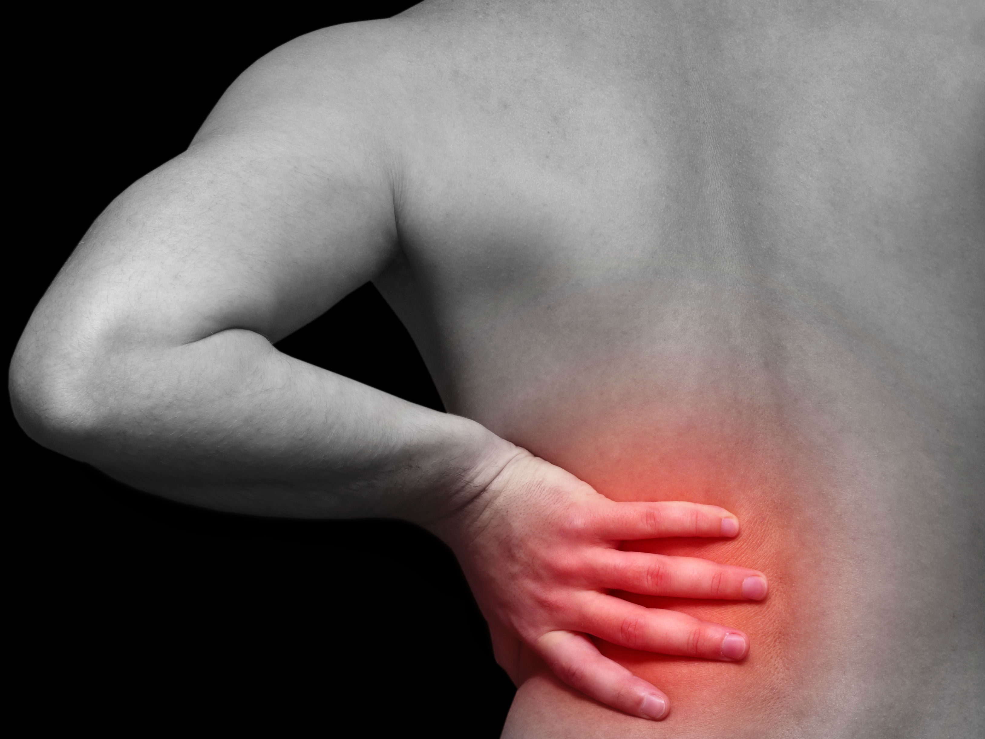 The image shows a close-up of a person’s back, highlighting a hand placed on the lower back, indicating pain or discomfort, with a dark background.