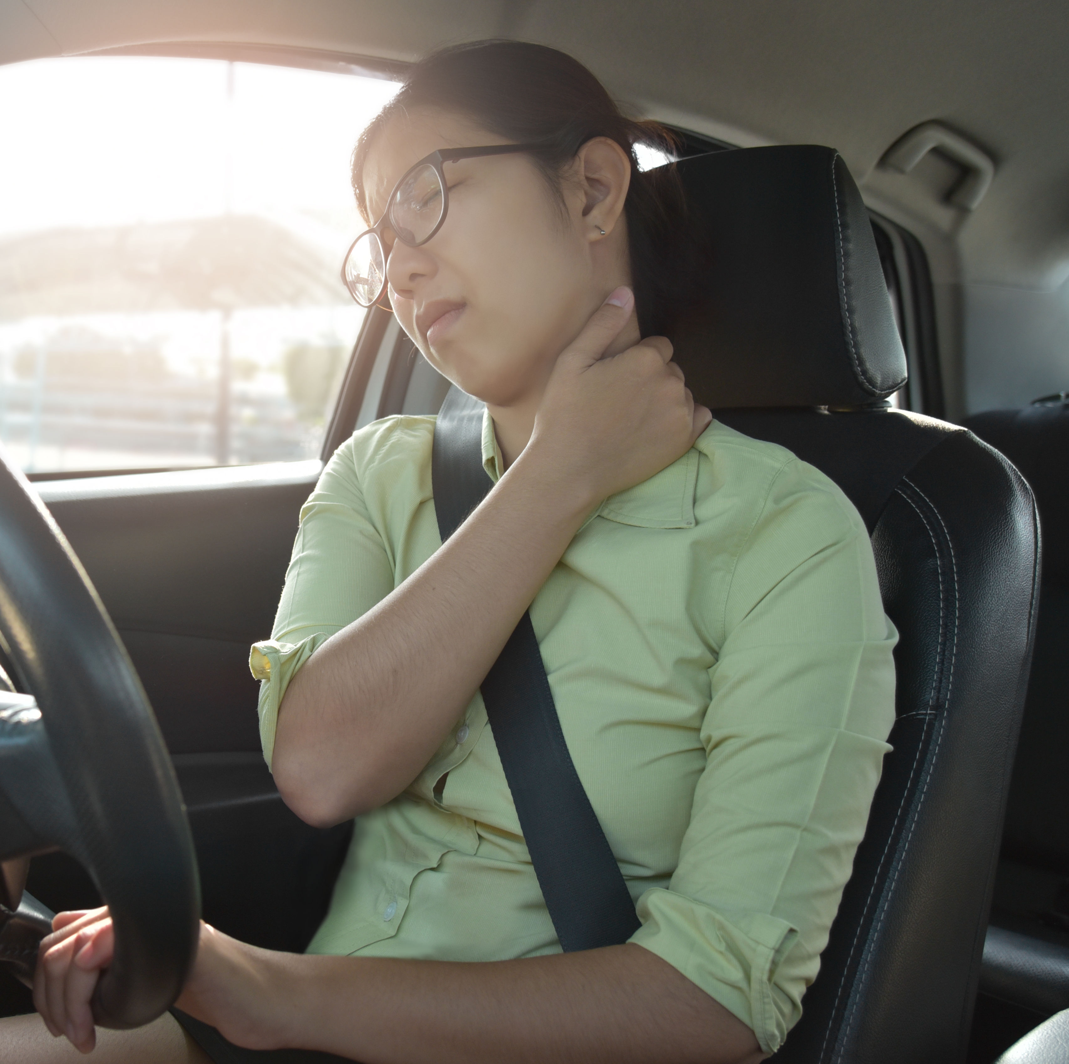 A woman sitting in a car grips the steering wheel, appearing to stretch her neck in discomfort, with sunlight streaming through the window.