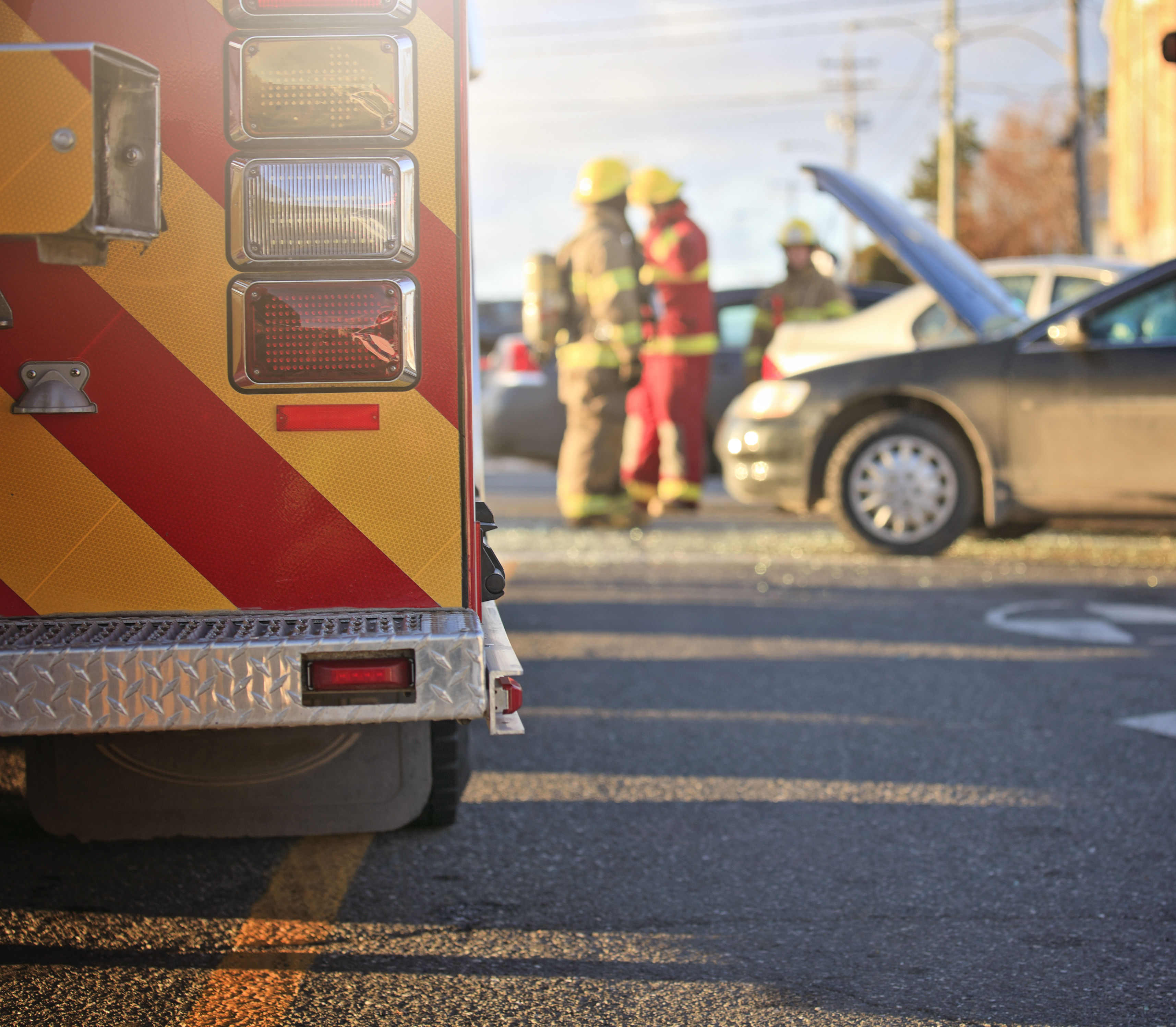 The image shows a fire truck with flashing lights, firefighters attending to a car accident, and a car with its hood raised on a city street.