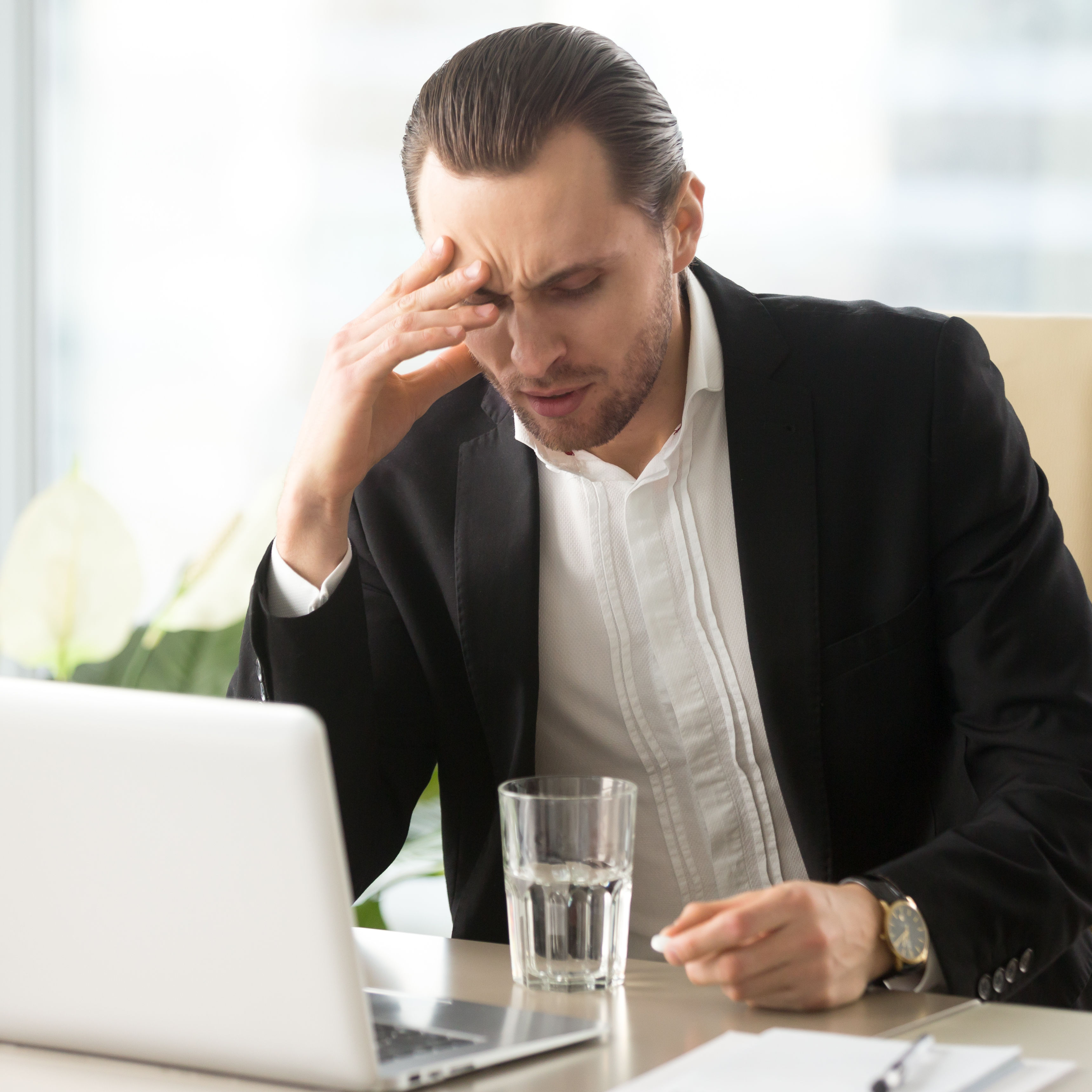 A man in a suit sits at a desk with a laptop, showing signs of stress or concern, while holding his head with one hand. A glass of water is nearby.