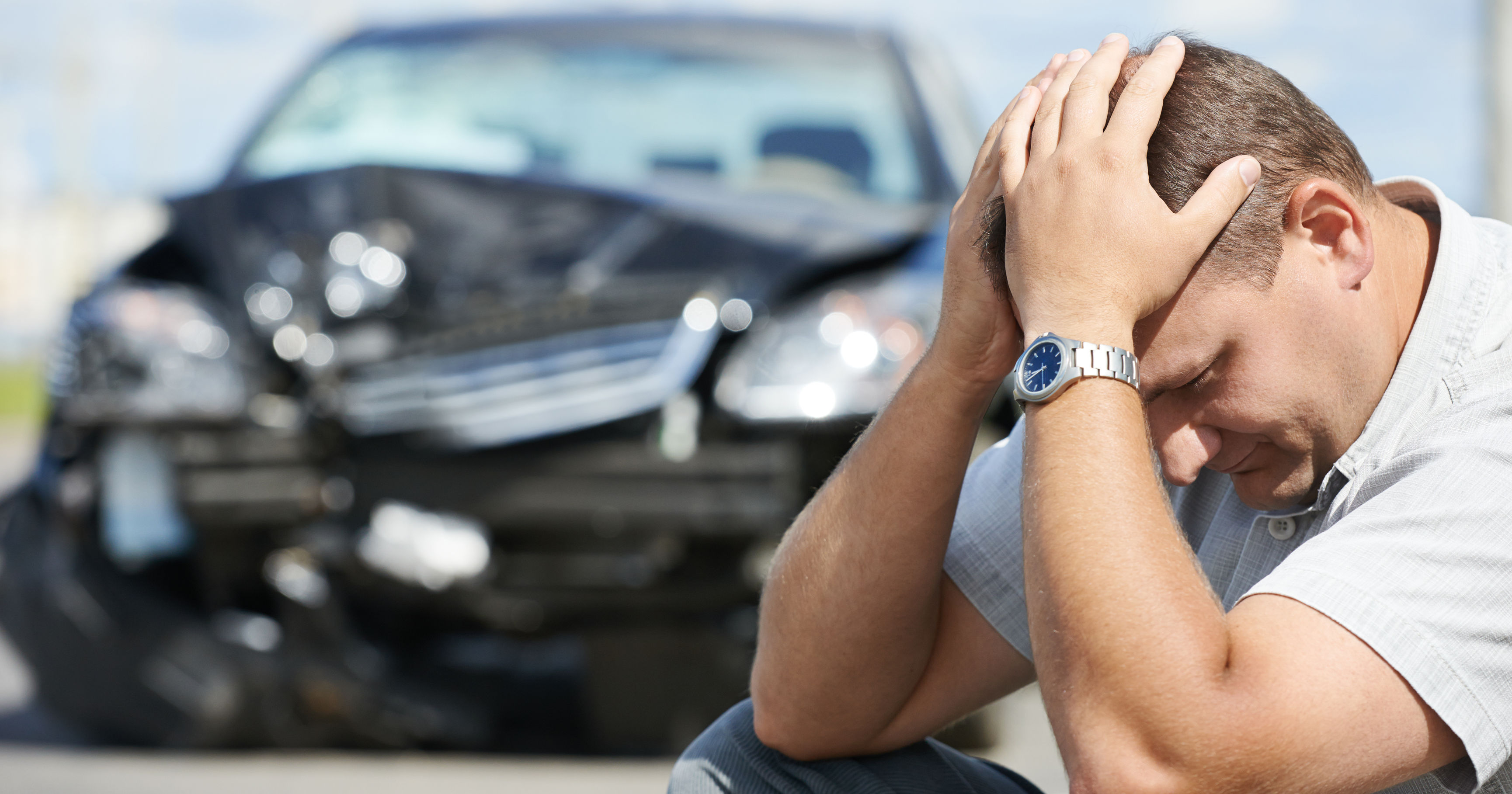 A man sits on the ground looking distressed, holding his head, with a damaged black car in the background, indicating a recent accident.