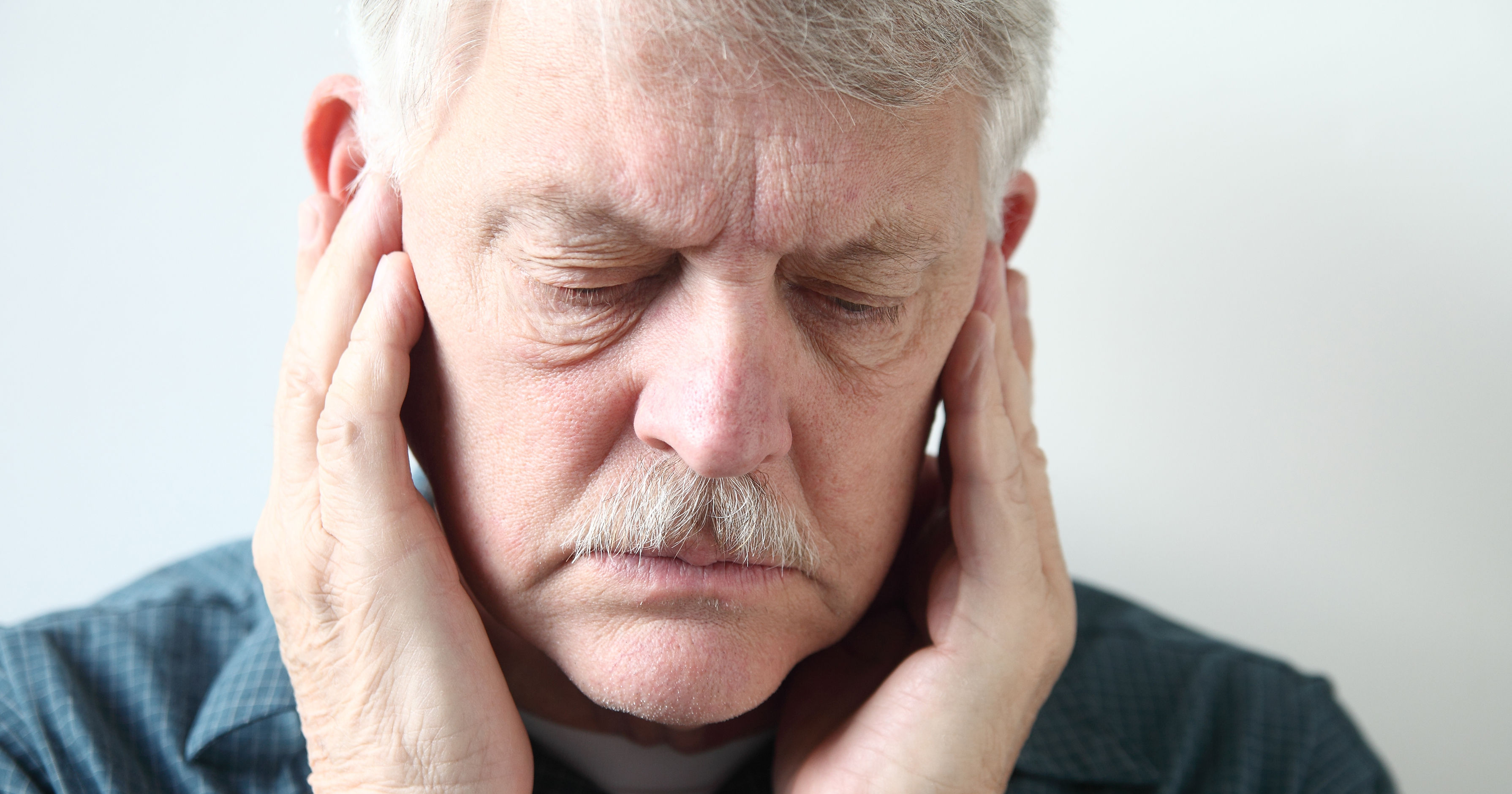An elderly man is holding his head with both hands, eyes closed, appearing distressed or overwhelmed.