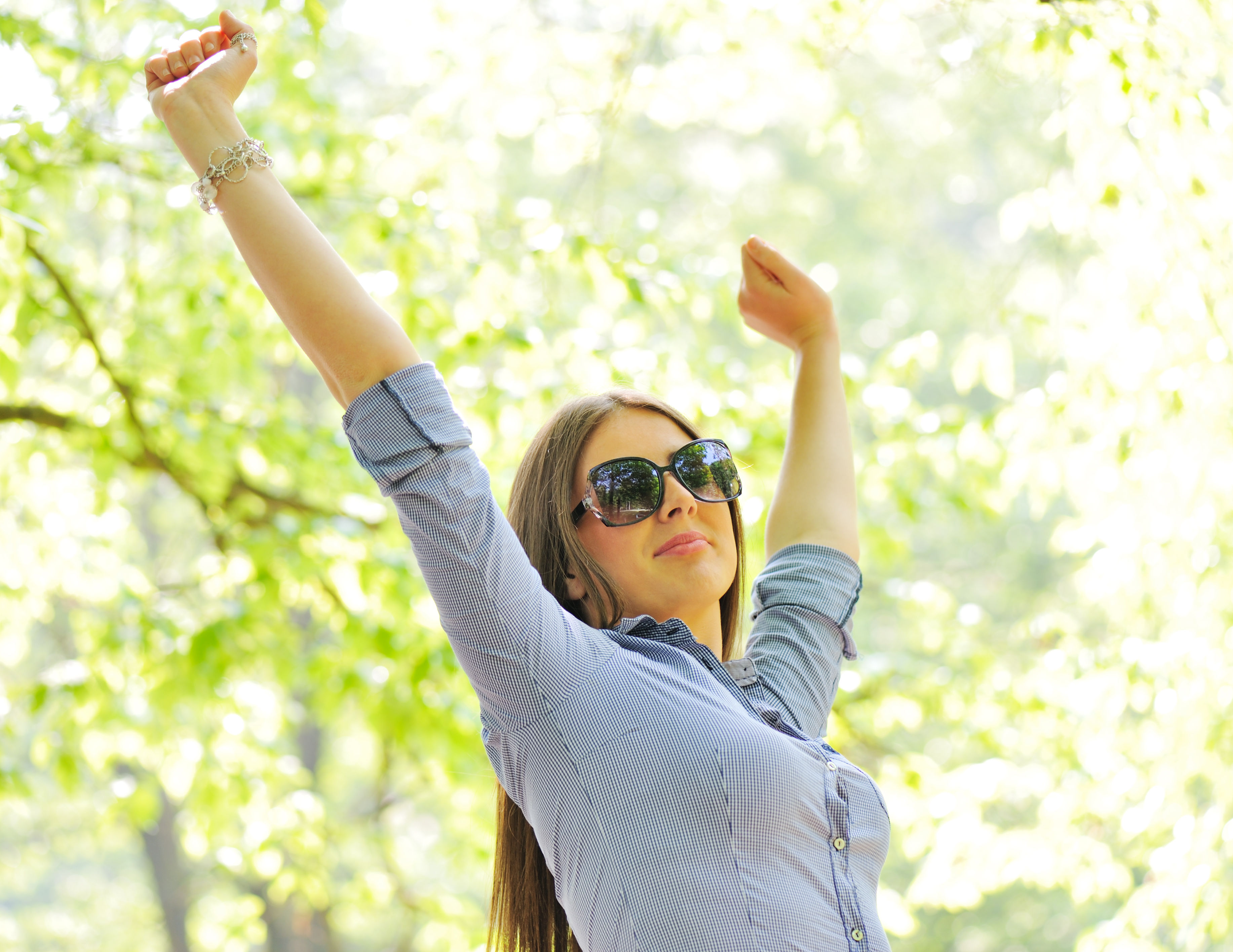 A woman with long hair raises her arms in a joyful pose, wearing sunglasses and a button-up shirt, amidst a bright, leafy background.