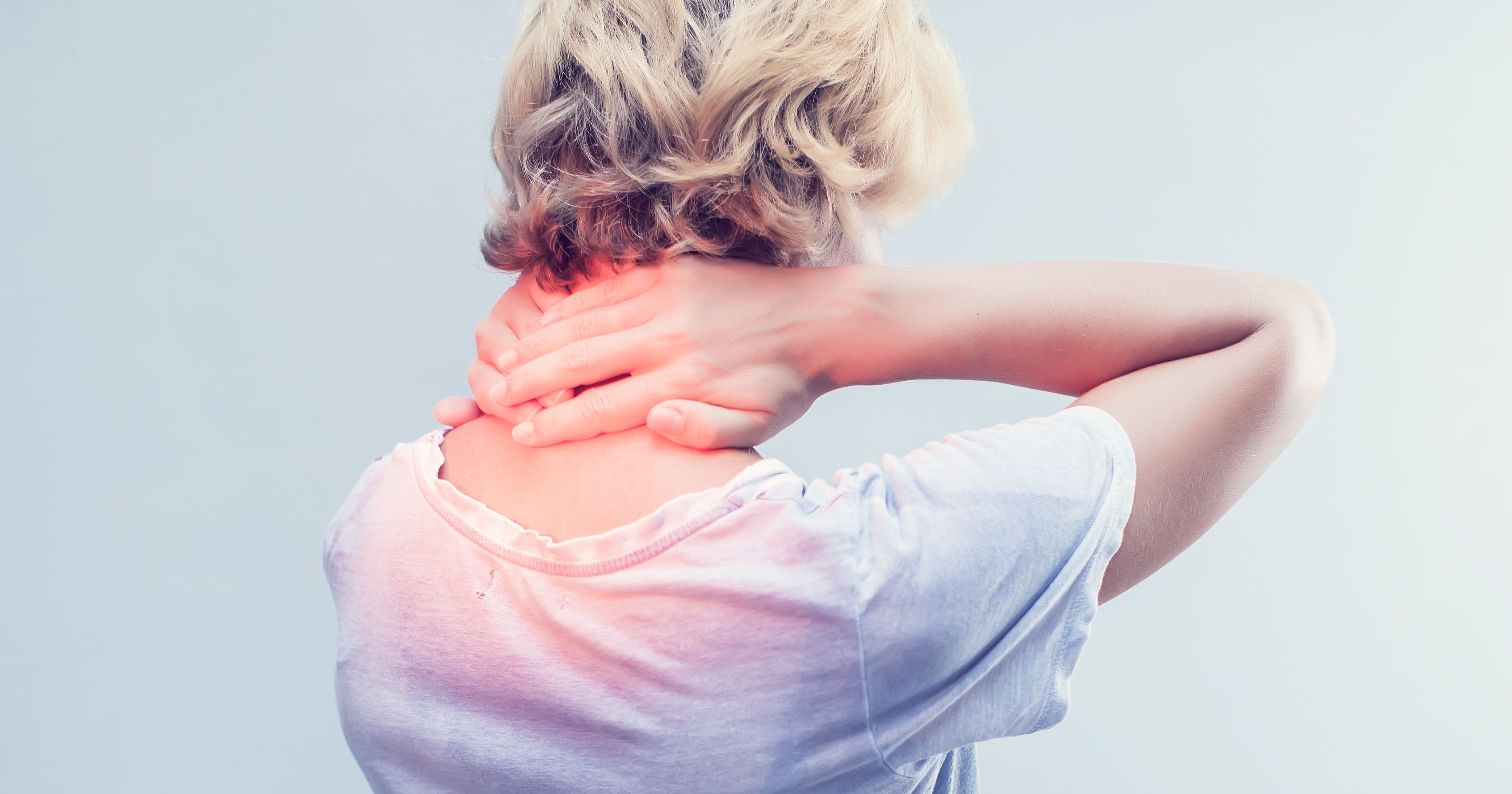 A person with short hair is seen from behind, holding their neck in discomfort while wearing a loose-fitting white shirt against a light background.