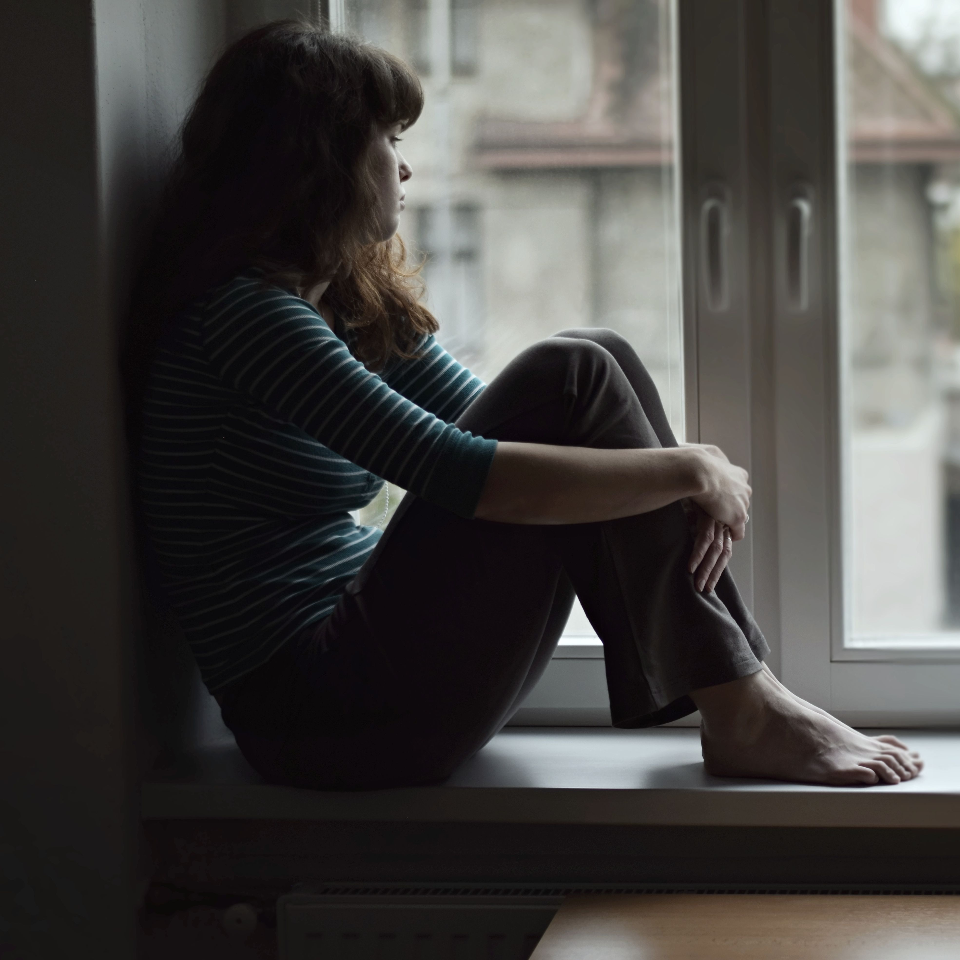 Sad young woman sitting on the window, watching ou
