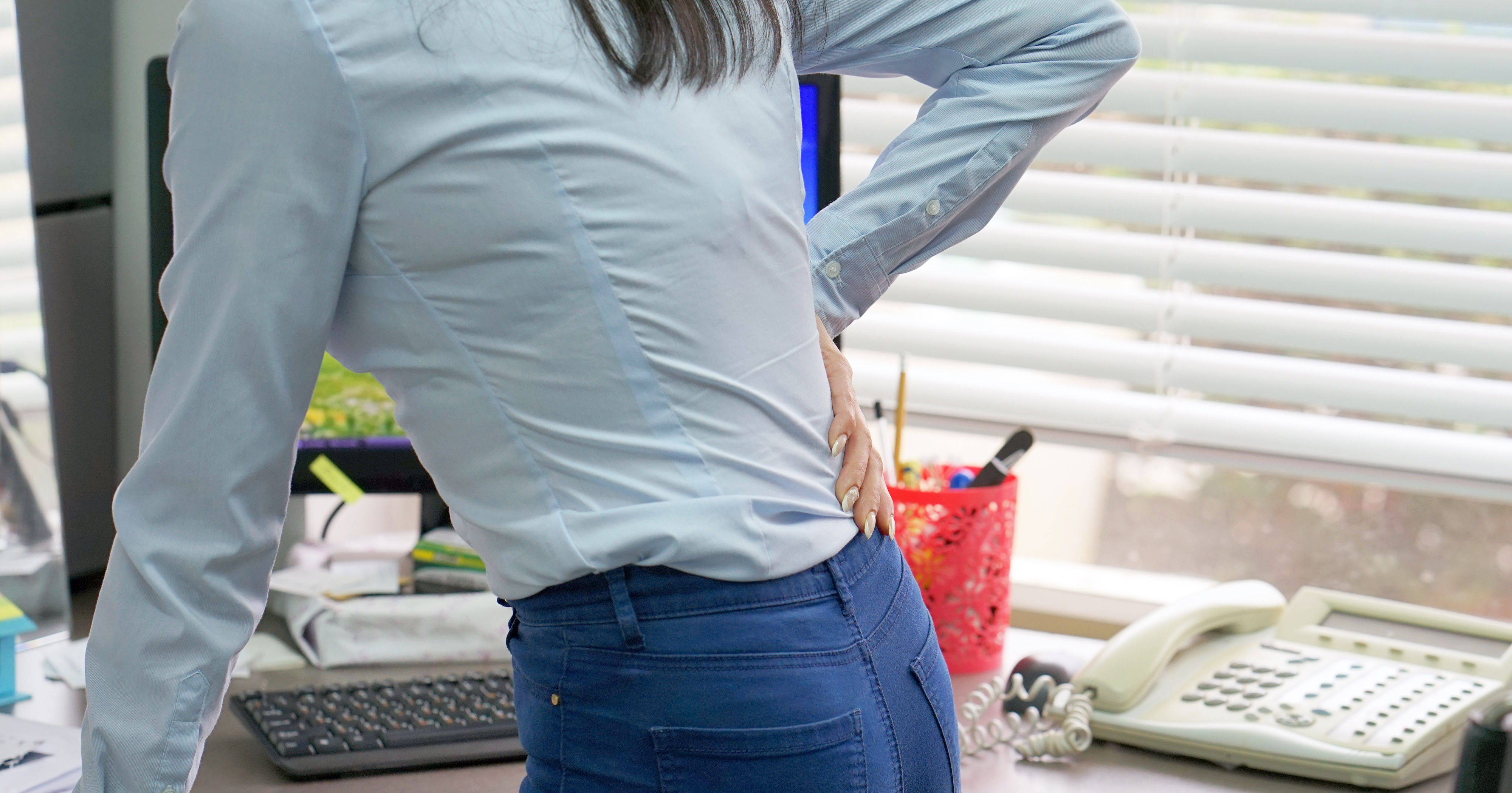 close up from the back of female office worker standing at desk holding low back pain