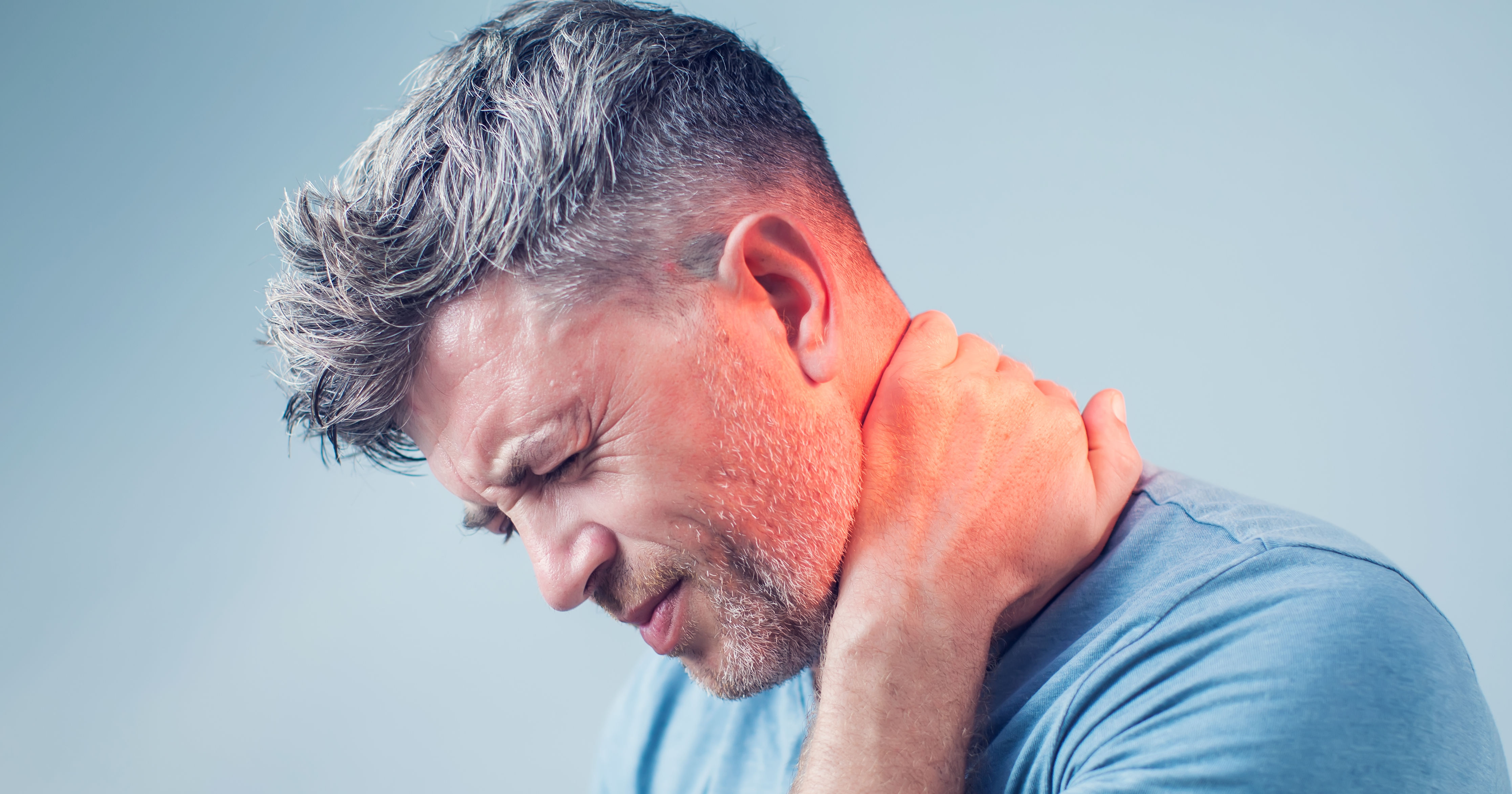 A man is holding his neck in discomfort, showing signs of pain or strain. He has short, styled hair and is wearing a blue shirt against a light background.