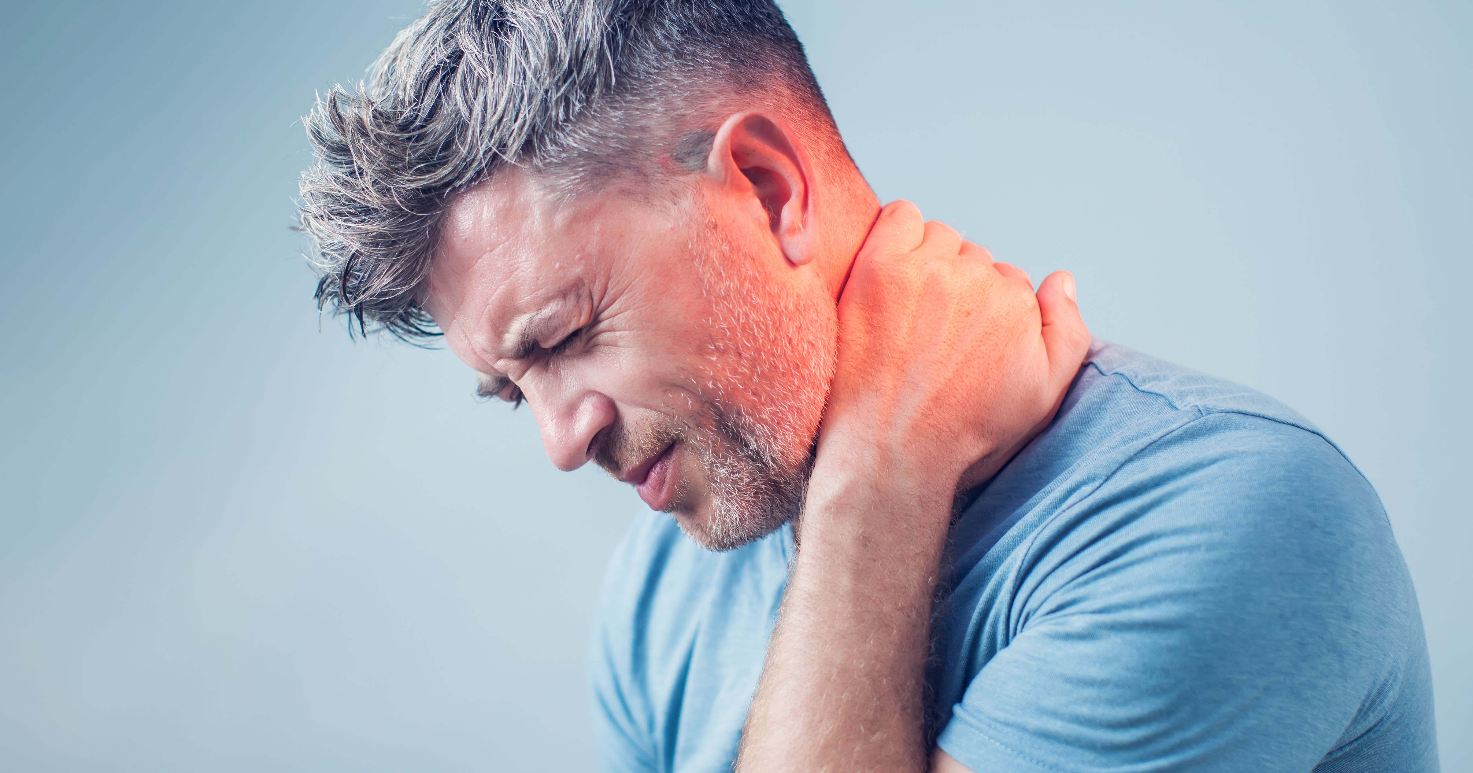 A man is holding his neck in discomfort, showing signs of pain or strain. He has short, styled hair and is wearing a blue shirt against a light background.