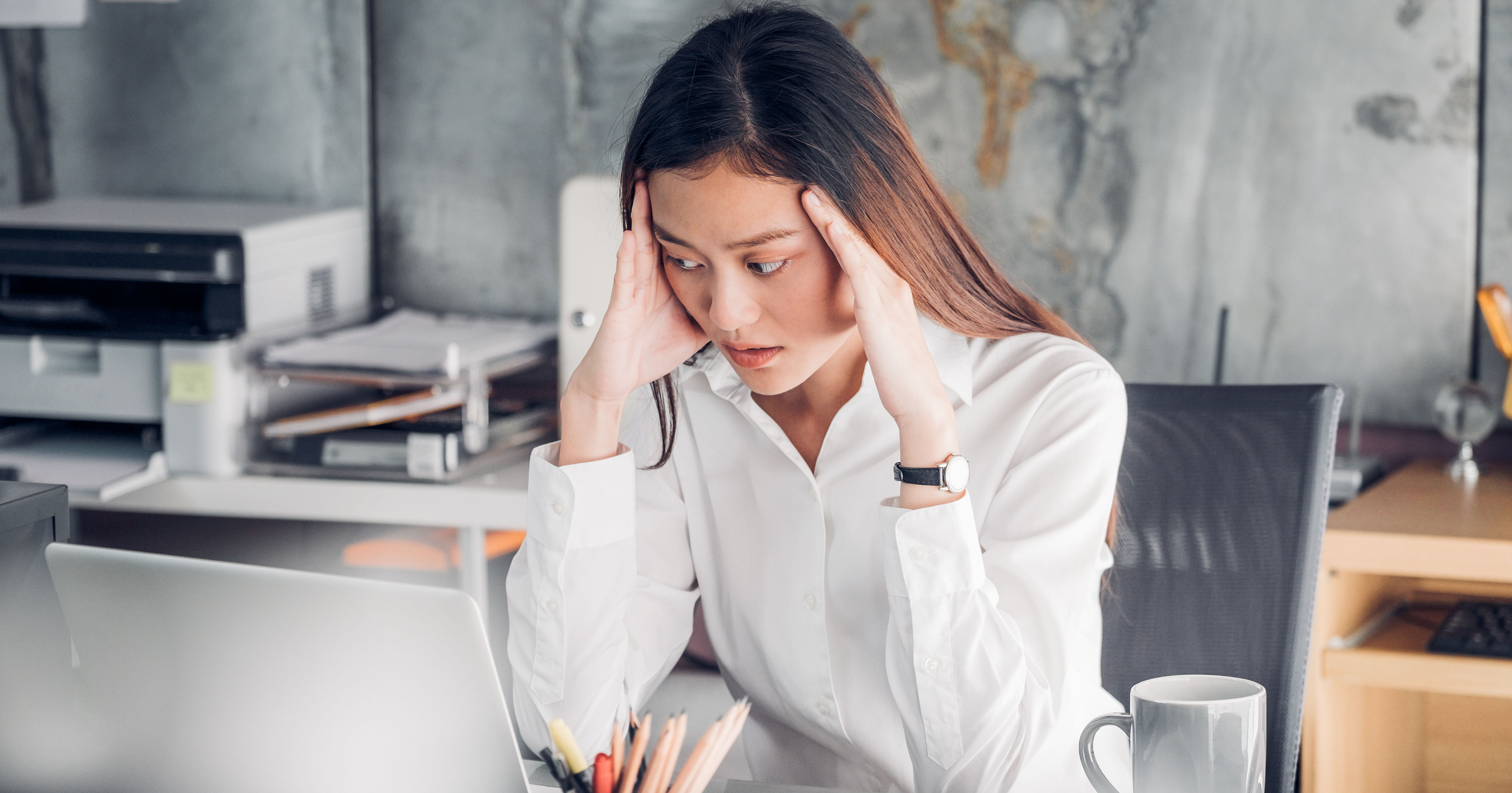 A woman in a white shirt sits at a desk, looking stressed with her hands on her head, surrounded by office supplies and a laptop.
