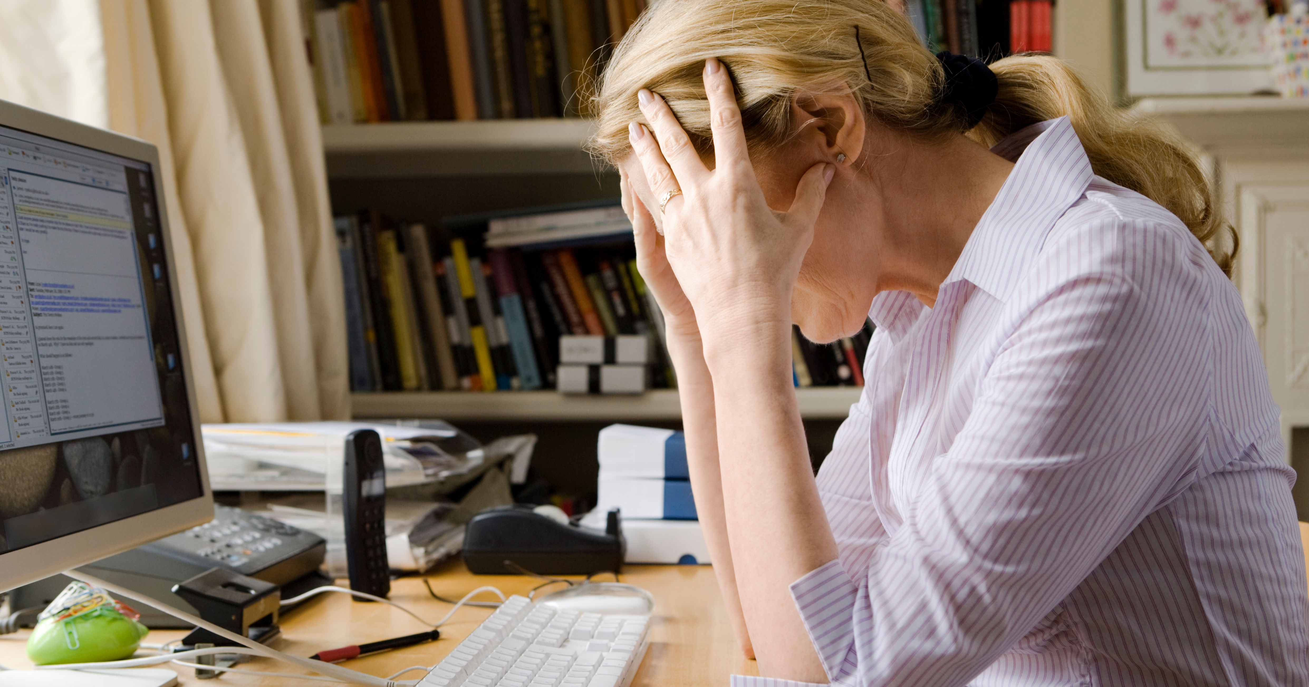 A person sits at a desk with a computer, looking stressed and frustrated, amid papers and a cluttered workspace.