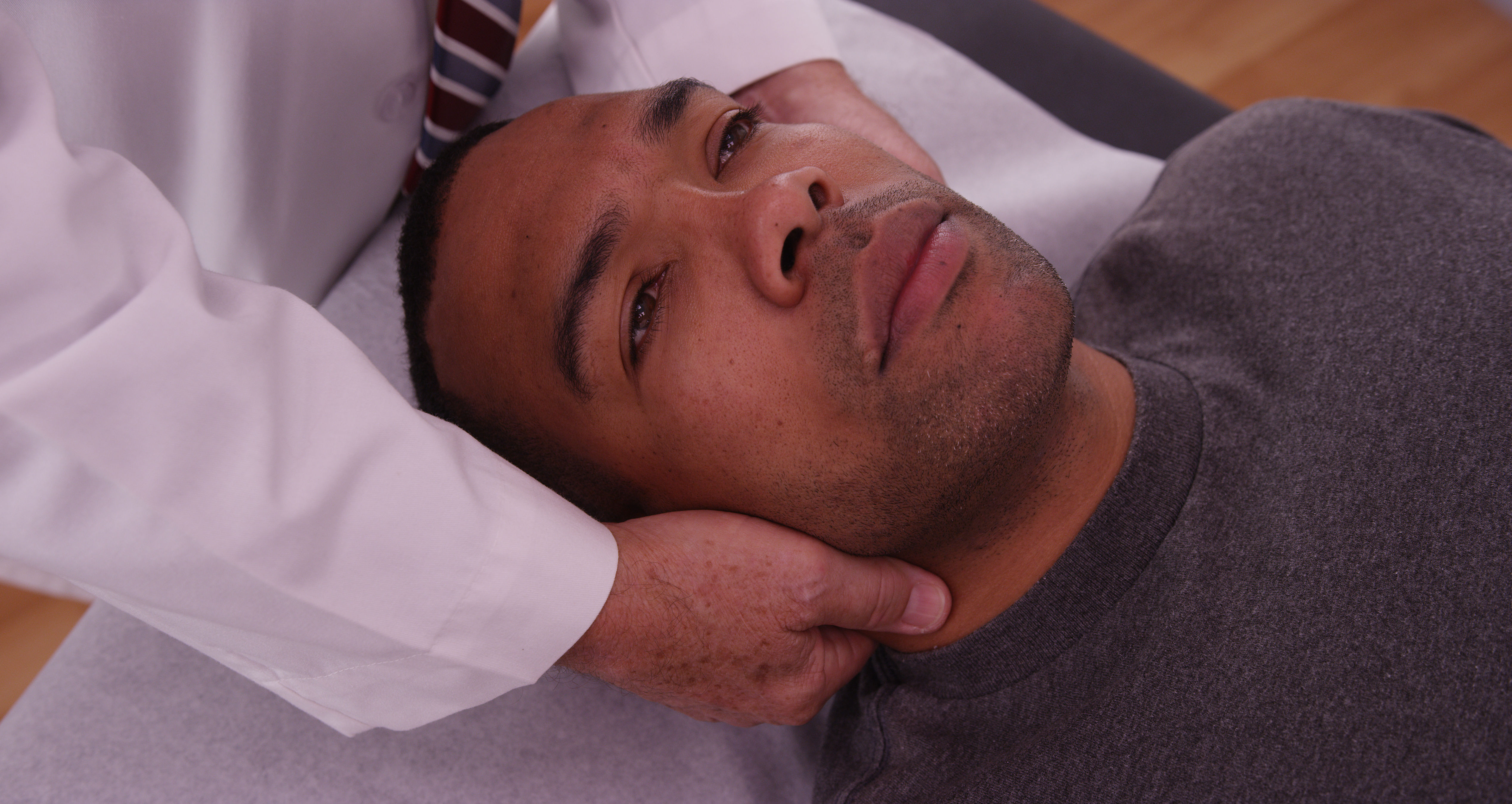 The image shows a man lying on a treatment table while a person in a white coat gently holds his head, possibly during a medical examination or therapy.