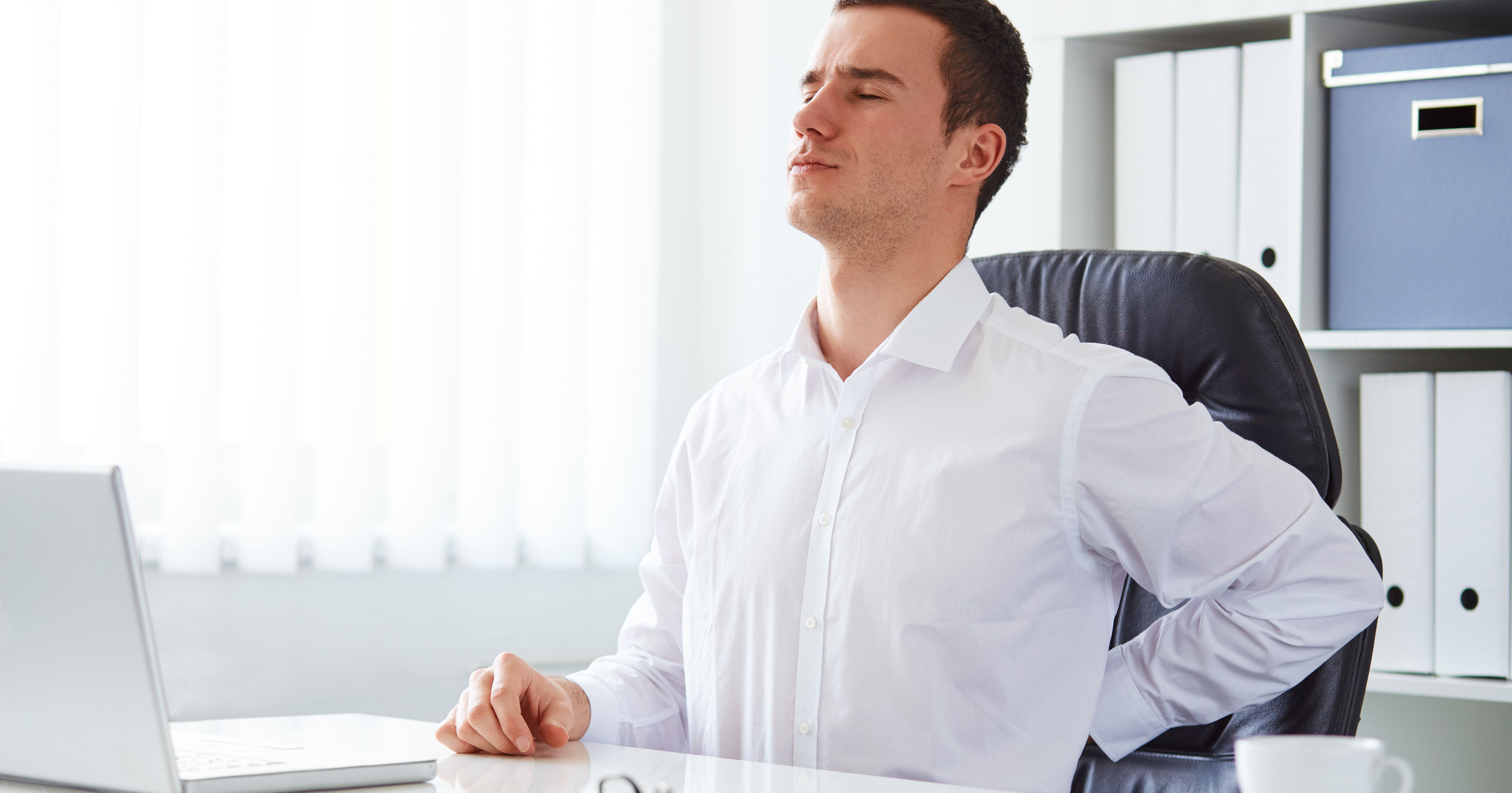 A man in a white shirt sits at a desk, leaning back in a chair with a pained expression, likely due to back discomfort, with a laptop in front of him.
