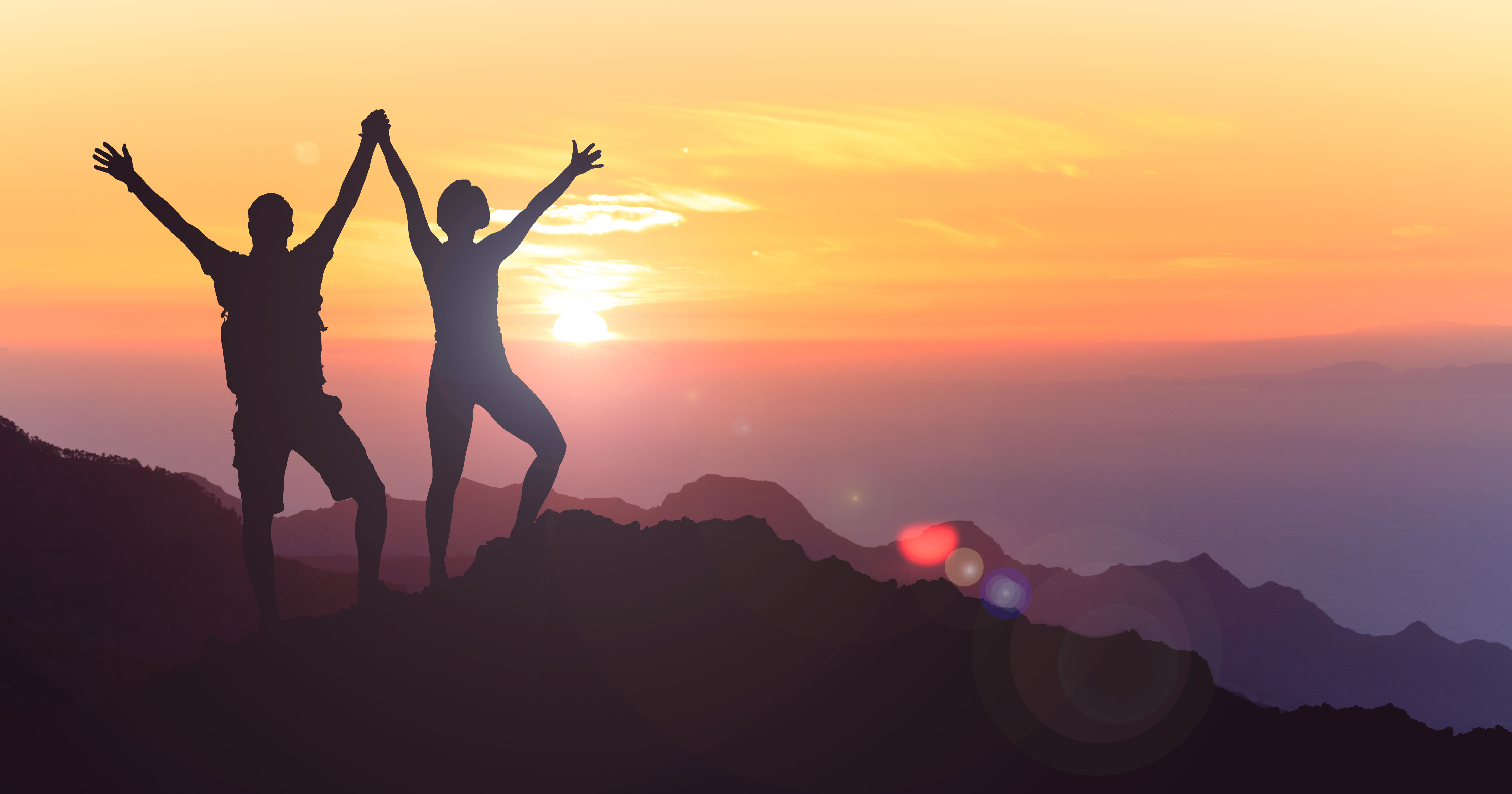 The image depicts two people silhouetted on a mountain peak, raising their arms in celebration against a vibrant sunset sky.