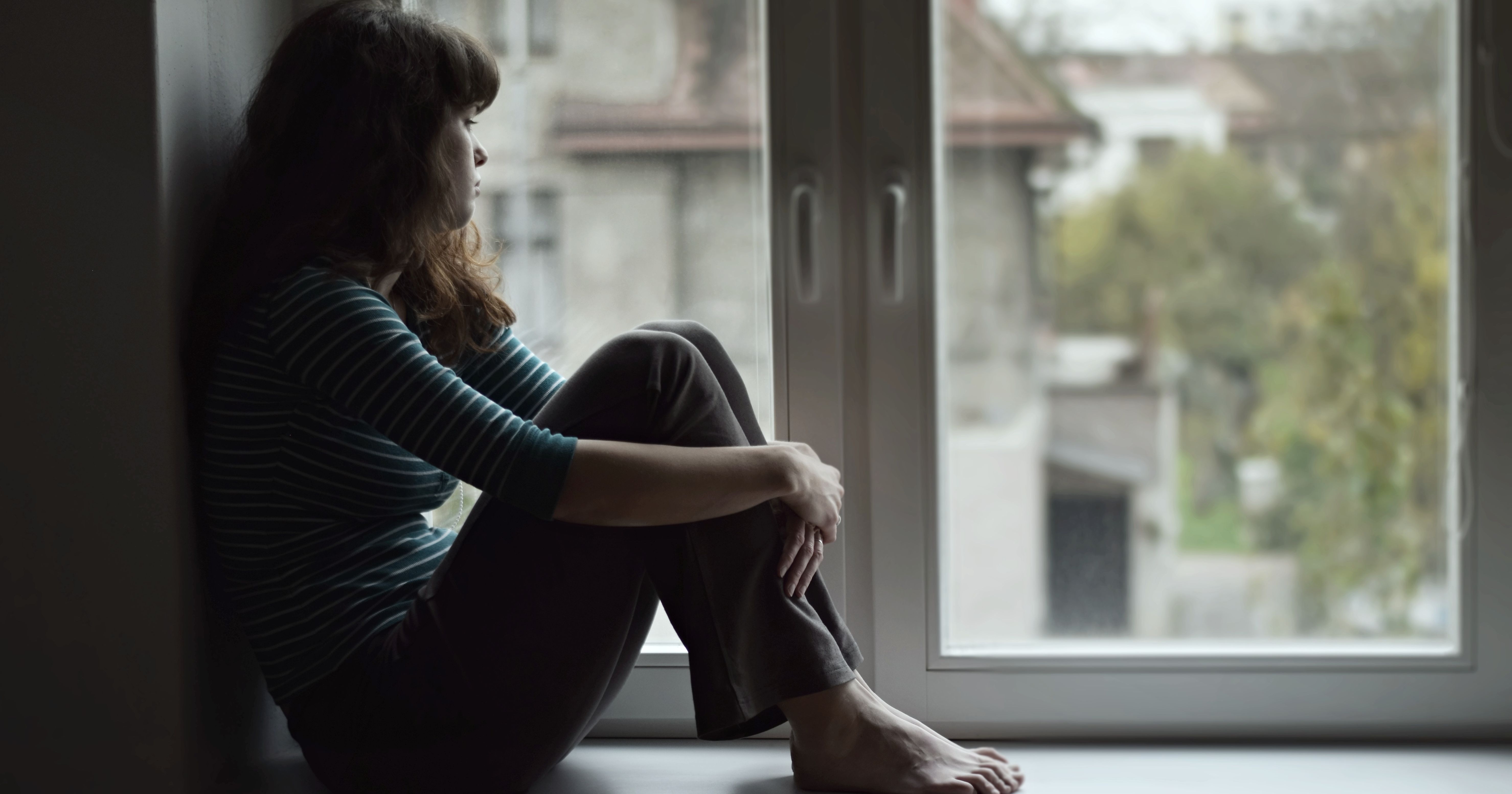 Sad young woman sitting on the window, watching ou