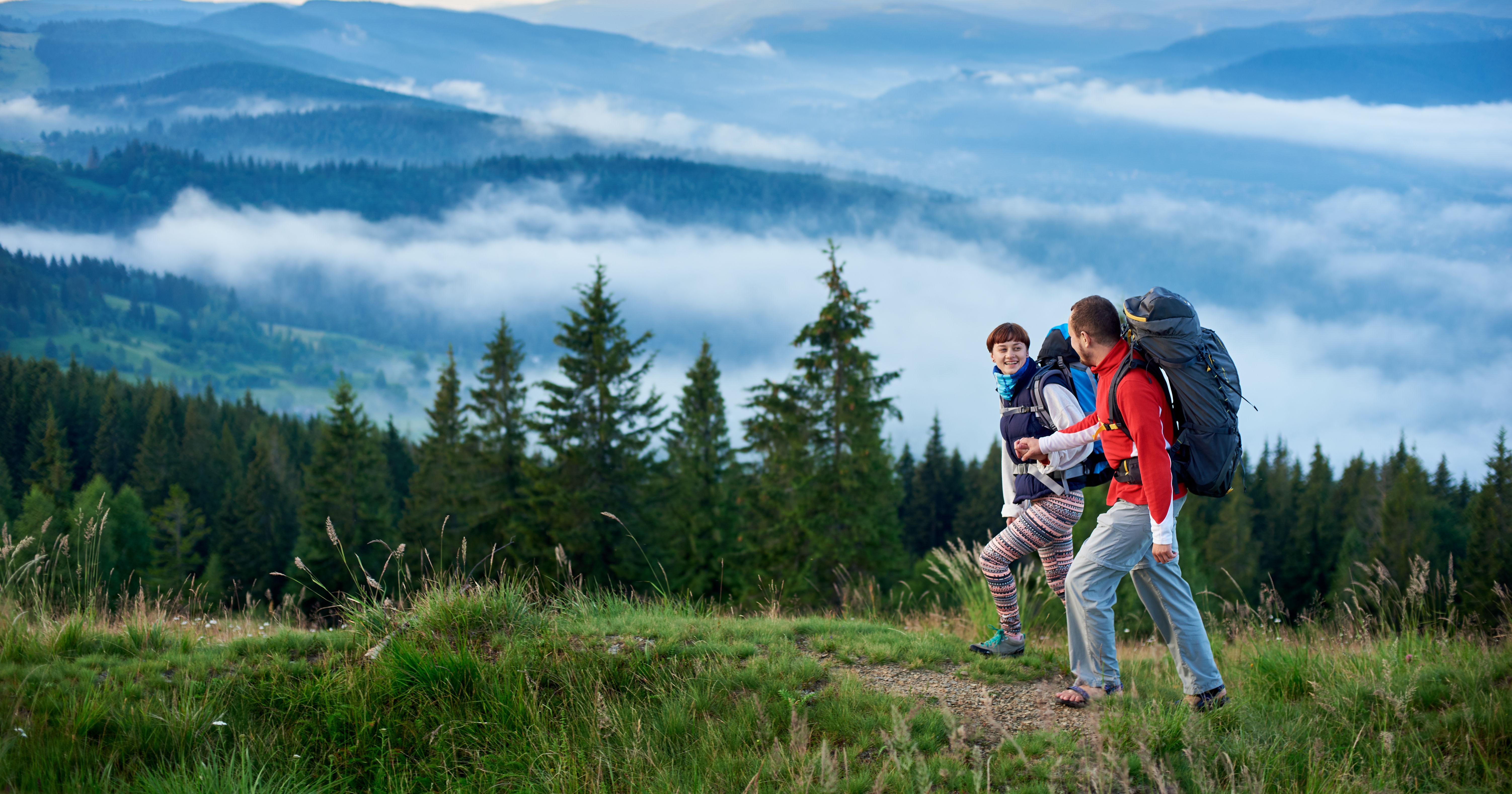 Two hikers are walking on a trail surrounded by lush greenery and misty mountains, enjoying the scenic view.