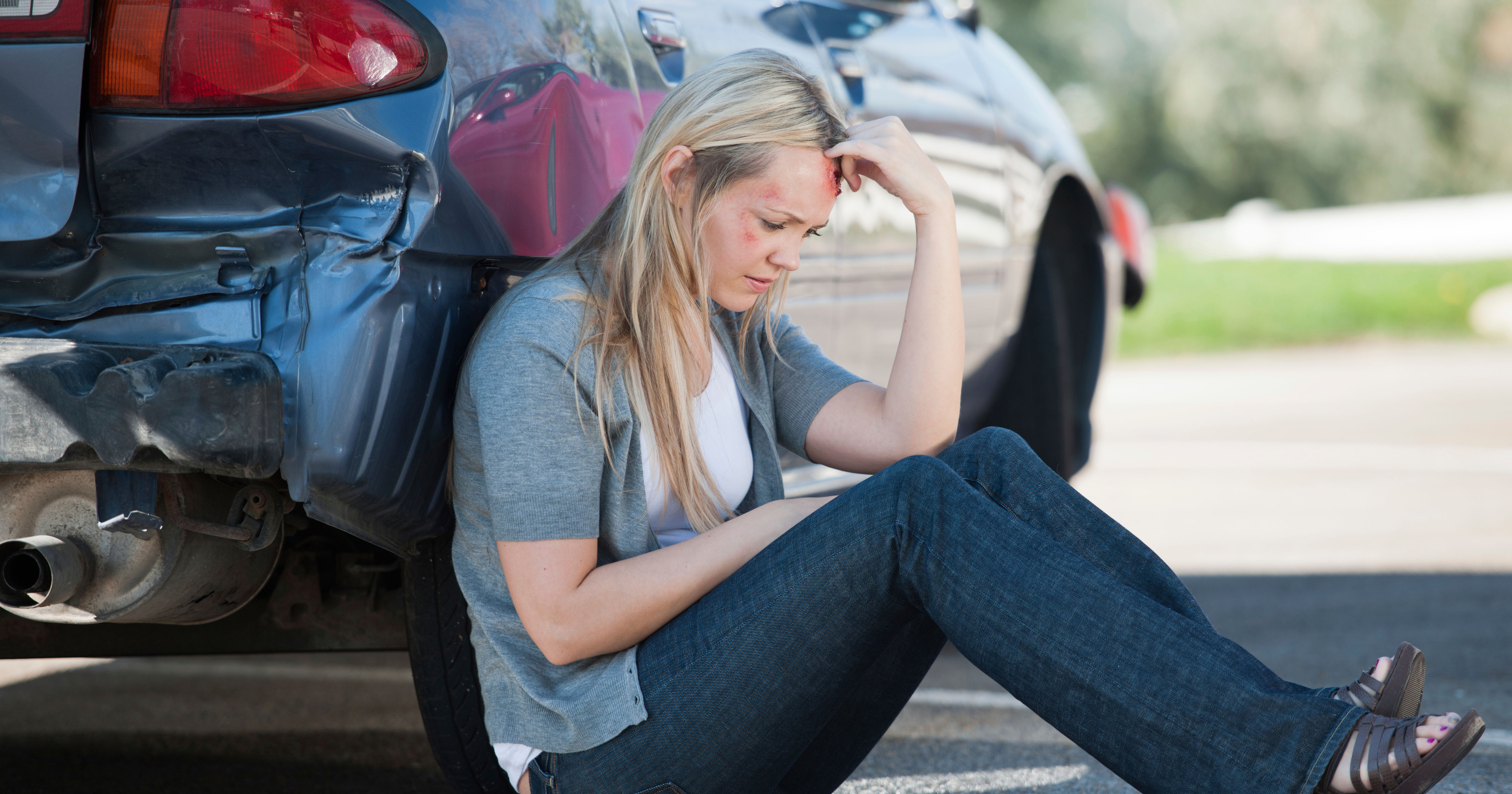injured person sits on ground beside damaged car