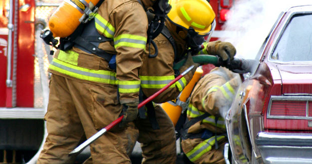 Firefighters in protective gear are extinguishing a fire in a car while a fire truck is visible in the background, with smoke rising from the vehicle.