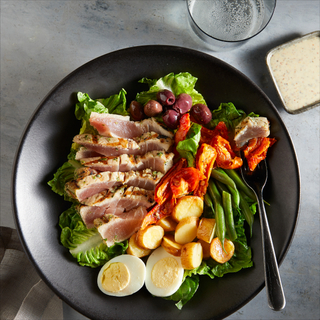 Ahi tuna niçoise salad with greens, sliced rare tuna, hard boiled eggs, steamed potatoes, olives in a black bowl with a fork on a grey background. 