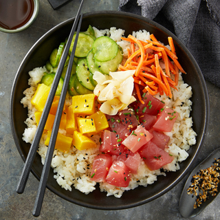 Black dinner bowl filled with white sushi rice, ahi tuna poke in cubes, matchstick cut carrots, sliced cucumbers, cubed mango, pickled ginger with black chopsticks resting on top and a grey napkin on the side. 