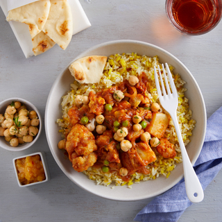 White bowl of aloo gobi on yellow rice with naan on a light surface, glass of wine and condiments visible 