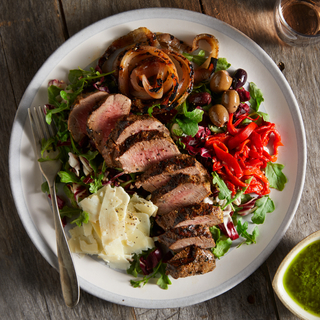 Round white plate filled with sliced Argentinian steak on a bed of greens with olives and shaved manchego cheese with a silver fork resting on top. 