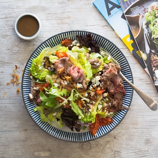 Striped blue rimmed bowl with greens and tri tip with a fork, magazines shown in the background.