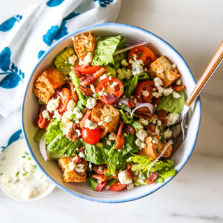 Bacon and blue cheese salad with sliced tomatoes, lettuce, crumbled bacon and blue cheese dressing in a white and blue bowl on a white marble background with a blue and white napkin