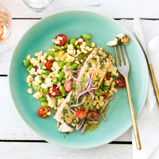 Teal bowl of basil roasted salmon over a salad of couscous, tomatoes and greens with a bottle of red wine and two glasses on a white wooden tabletop. 