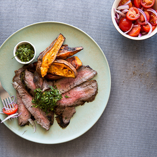 sliced steak with green chimichurri sauce and roasted sweet potatoes and a side tomato salad