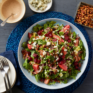 Blue dinner plate with beets and blue cheese salad on a dark blue placemat with dressing on the side, a dish of blue cheese crumbles and two small plates with forks on top. 