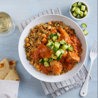 Butter chicken curry over confetti quinoa with cucumber pickles on top in a white bowl with a side of naan wrapped in paper on a striped napkin with a fork and a glass of white wine on the side. 