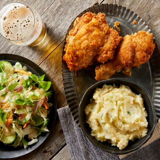 Buttermilk fried chicken drumstick and thigh on a tin tray with mashed potatoes and a salad on the side with a glass of beer on a wooden table. 