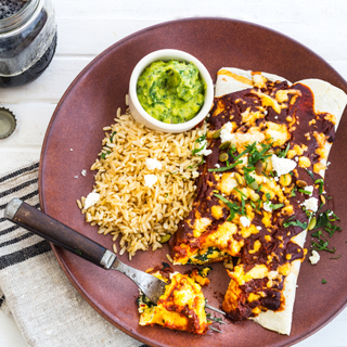 Butternut squash enchiladas smothered in sauce and cheese with rice and guacamole in a round dish on the side of a dark red plate with a linen napkin underneath. 