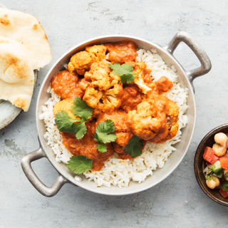 Cauliflower tikka masala over white basmati rice in a pot with handles next to a plate with naan and a bowl of relish on the side. 