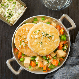 Silver dish with handles filled with chicken and biscuits topped with fresh herbs on a wooden table with a grey napkin. 