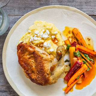 Roasted chicken thigh with skin on over feta polenta with cooked carrots on a white plate on a wooden table. 