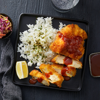 Crispy chicken katsu sliced and covered in hot sauce on a bed of white rice with a fresh lime wedge on a black square plate on a dark wooden background. 