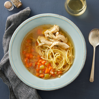 Chicken noodle soup with spaghetti noodles and vegetables in broth in a white bowl on a dark blue background with a spoon and a glass of white wine. 