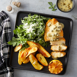 Chicken piccata breast with caper sauce next to fresh arugula salad with cheese and roasted sliced potatoes and a charred lemon half on a black square plate. 