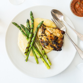 Grilled chicken thigh with creamy polenta and charred asparagus on a white plate with a fork and spoon on the side with a glass of white wine and a side of red sauce. 