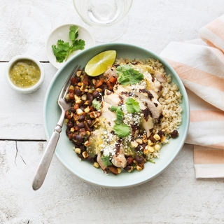 Blue bowl on a white wooden table with chicken verde rice bowl with fresh lime, beans, corn, brown rice and chicken thighs