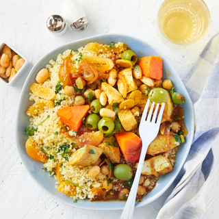 Chicken tagine with green olives, dried apricots, almonds and rice in a blue bowl with a fork and a glass of white wine on the side. 