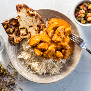 Chicken tikka masala on white basmati rice with crisp naan and a side of vegetables with a fork on a white background. 