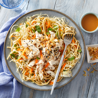 Chinese chicken noodle salad on a grey plate with a silver fork resting on top with brown dressing on the side and a blue napkin.