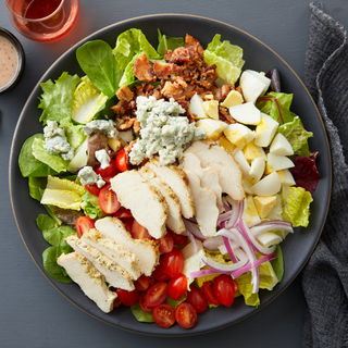 Cobb salad with sliced grilled chicken, crumbled blue cheese, chopped hard boiled eggs, cherry tomatoes, crumbled bacon and sliced red onion on chopped lettuce in a blue bowl on a blue background. 
